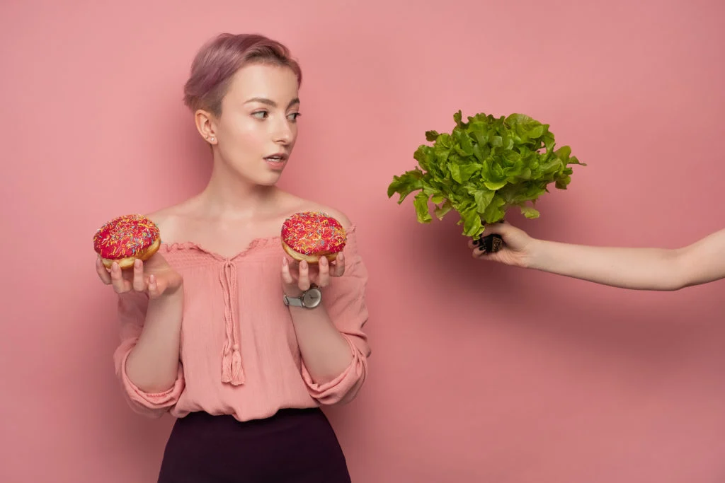 A woman holds two doughnuts, while a hand emerging from out of frame offers her a head of romaine lettuce.