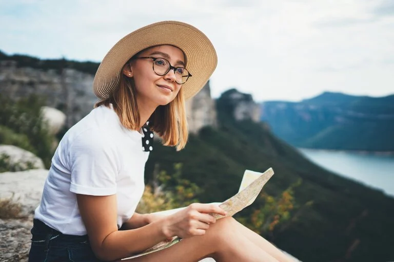 A woman wearing glasses and a sunhat sits at a scenic cliffside.