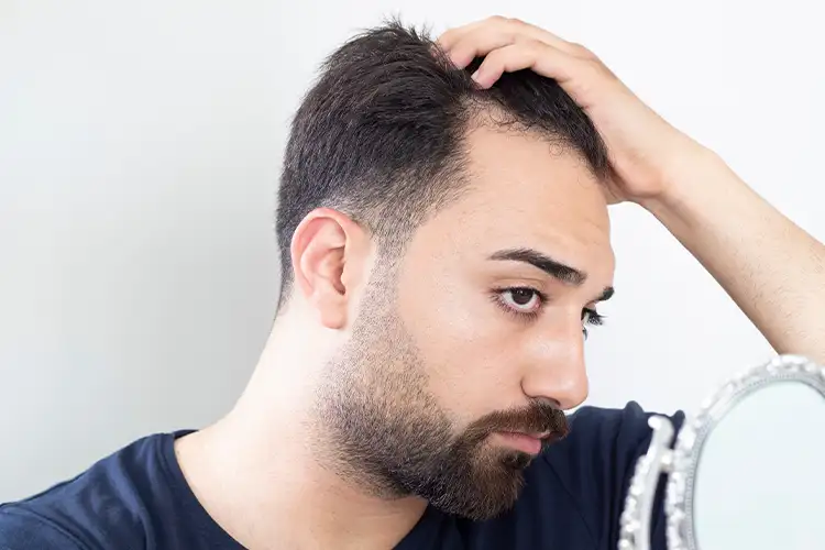 A man looks thoughtfully at his hairline in the mirror – with an uncertain look and visible doubt about his appearance