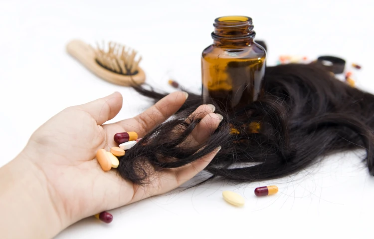 A woman holds several pills and some of her lost hair in her hand, with more pills, a medication bottle, strands of hair, and a brush in the background.