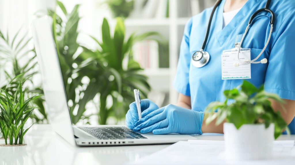 A doctor wearing blue scrubs and blue surgical gloves types at a laptop surrounded by plants.