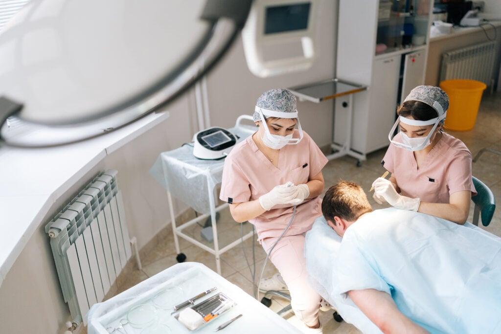 Two female medical technicians perform a follicular unit extraction on a male patient.