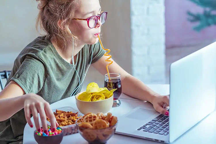 A young woman studies at a laptop, eating a variety of junk food snacks.