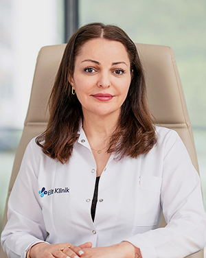 Elithair's senior dermatologist, Dr. Ghazaleh Jafari wearing a white coat with the logo “Elit Klinik,” seated and smiling slightly in a bright medical office.