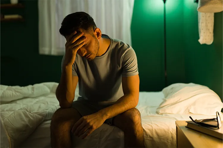 A distraught man sits on the edge of his bed.