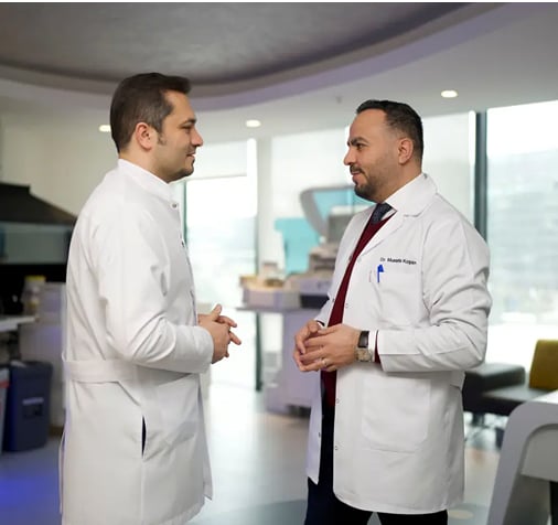 Doctor Balwi and doctor Moustafa standing in Elithair's research area, engaged in conversation with medical equipment in the background.
