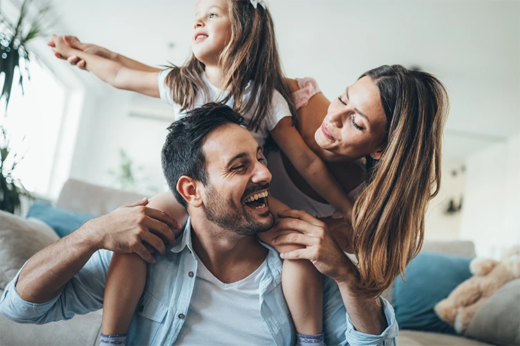A man holds his daughter on his shoulders, while his wife leans over towards them. All are smiling.