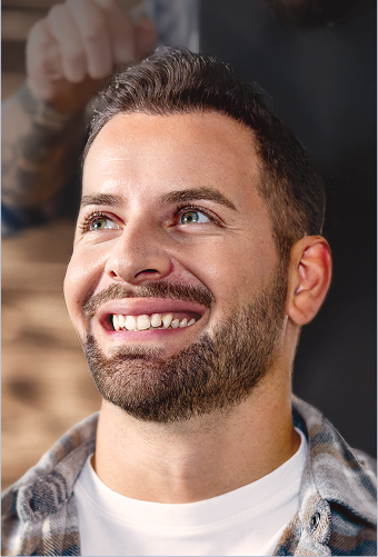 Smiling man with short dark hair and beard looking upward, wearing a white shirt and checked overshirt indoors.