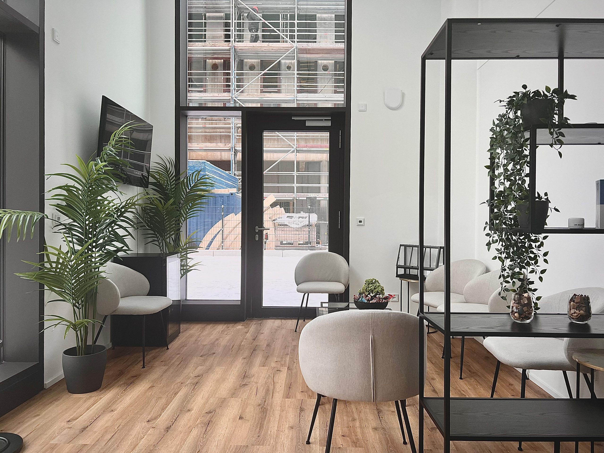 Modern waiting area with light chairs, wooden flooring, indoor plants, and large glass doors showing a building under construction outside.