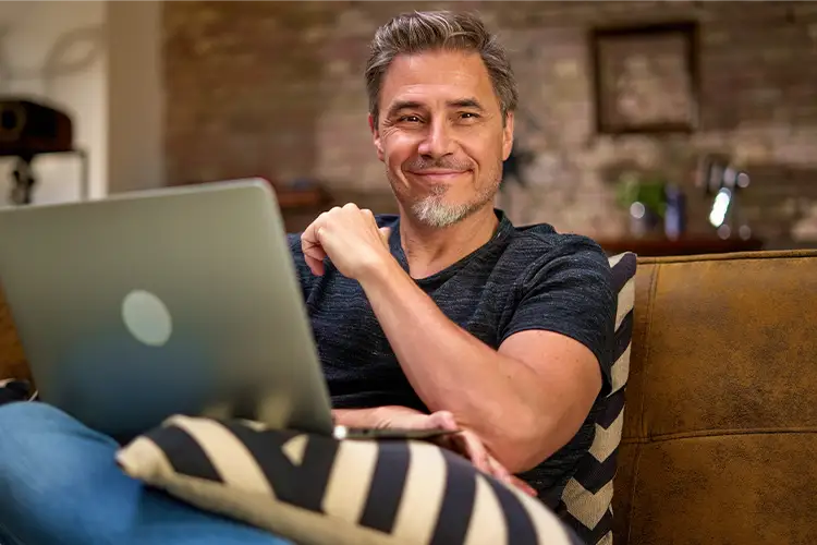 A man with a full head of hair sits on a sofa with a laptop on his lap, smiling at the camera.