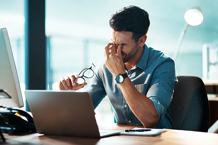 A frustrated man at a laptop pinches the bridge of his nose.