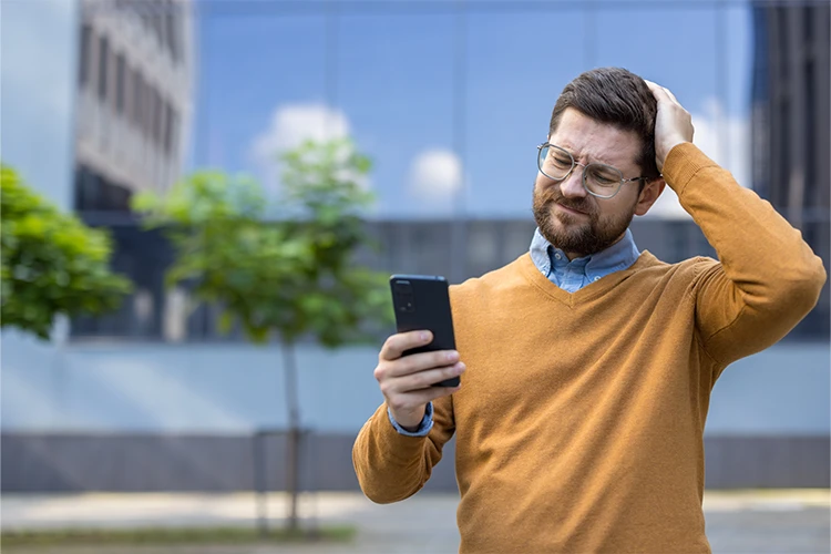 A man rubs his head in confusion while looking at his smart phone.