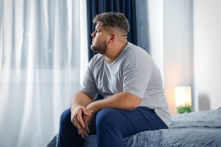 An overweight man sits on the edge of a bed, looking pensive.