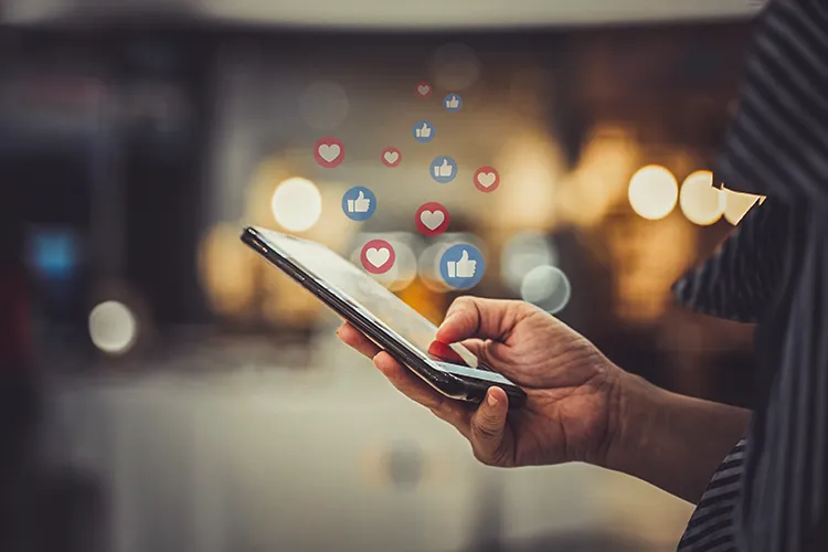 A hand holding a smartphone, from which illustrated depictions of likes and other social media engagements flow.