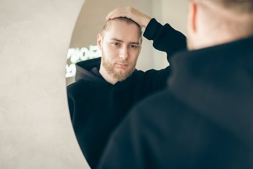 A young man examines his hairline in a mirror.
